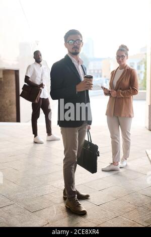 Porträt eines seriösen bärtigen Business-Spezialisten in Brillen mit Kaffeetasse und Aktentasche, seine Kollegen hinter ihm stehen Stockfoto