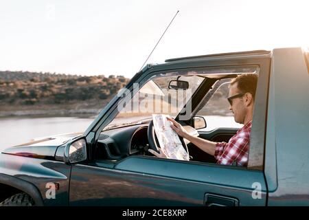 Seitenansicht des anonymen männlichen Reisenden in legerer Kleidung und Sonnenbrillen beobachten Karte, während im Auto vor sitzen Fluss und Berge unter ruhigem Himmel Stockfoto