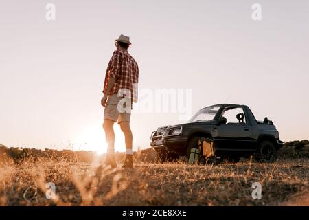 Rückansicht von anonymen männlichen Touristen in Freizeitkleidung und hut Genießen Sie die malerische Natur, während Sie auf verblassenem Gras in der Nähe stehen auto unter heiterem Himmel bei Sonnenschein Stockfoto