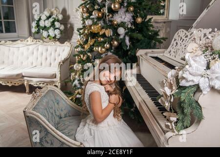 Frohe Weihnachten Happy Holidays Konzept. Nettes kleines Mädchen in weißem Kleid spielen auf Klavier vor dem Hintergrund eines verzierten Weihnachtsbaum. Festlich Stockfoto