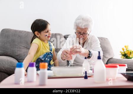 Lächelnde Oma in Brillen hilft positive Enkelin mit schmutzigem Gesicht Bei Freizeitkleidung Vorbereitung Mischung mit Farbstoffen in Kunststoffbehälter Während Sie auf einer bequemen Couch im Wohnzimmer sitzen Stockfoto