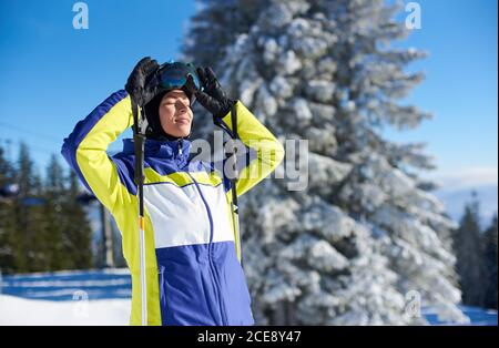 Porträt einer lächelnden Frau mit Skistöcken schließen ihre Augen vor strahlendem Sonnenschein, nachdem sie ihre Brille entfernt hat. Skifahrer, die zum Ausruhen anhalten und die malerische Aussicht auf die Berge genießen. Zugeschnittenes Bild. Stockfoto