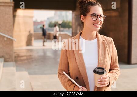 Positive erfolgreiche junge Geschäftsfrau in Brillen trinken Kaffee in Bewegung Beim Spaziergang über die Stadt Stockfoto
