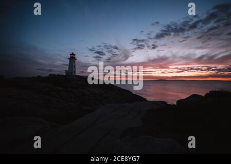 Peggys Cove Lighthouse liegt an steiniger Küste gegen Meer und wolkiger Sonnenuntergang Himmel am Abend in Kanada Stockfoto