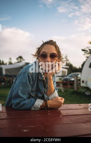 Friedliche weibliche Hippie sitzt am Holztisch auf dem Campingplatz und Ruhig wegschauen während der Sommerferien Stockfoto