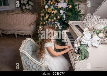 Frohe Weihnachten Happy Holidays Konzept. Nettes kleines Mädchen in weißem Kleid spielen auf Klavier vor dem Hintergrund eines verzierten Weihnachtsbaum. Festlich Stockfoto