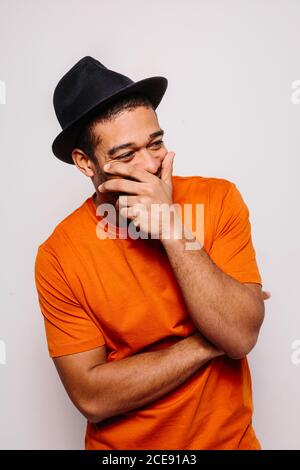 Fröhlicher Mann isoliert auf weißem Hintergrund. Er trägt ein orangefarbenes T-Shirt, einen schwarzen Hut und schaut mit der Hand auf dem Mund weg Stockfoto