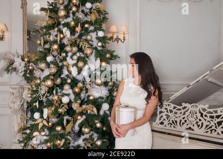 Frohe Weihnachten und frohe Feiertage. Schöne junge Brünette Frau in weißen Abend Cocktail-Kleid posiert mit einem weißen Klavier und geschmückt Weihnachten Stockfoto