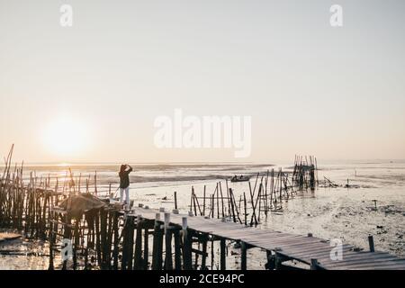 Blick aus der Ferne auf eine anonyme Frau, die auf einem alten Holzpier an der Küste mit flachem Wasser steht, Vicentina, Portugal Stockfoto