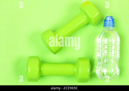 Paar grüne Fitness-Hanteln und eine Flasche Wasser auf grünem Hintergrund. Gewichtheben und Bodybuilding Konzept. Zwei Kunststoff-Kurzhanteln und klare Flasche. Set von Hand Gewichte und Wasser auf hellem Hintergrund. Stockfoto