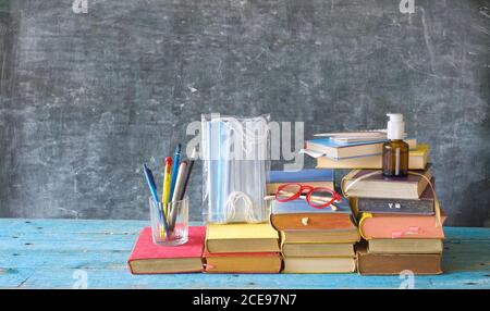 Schulbedarf und COVID 19 Vorbeugungsartikel auf Klassenzimmer Schreibtisch mit Büchern, Brillen, Stifte auf Kreidetafel Hintergrund. Zurück zur Schule während Corona-viru Stockfoto
