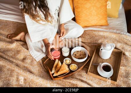 Gesundes Frühstück im Bett mit Müsli, knusprigem Toast, Butter, Milch und Marmelade. Frau Hand hält Wassermelone Saft auf Holztablett Stockfoto