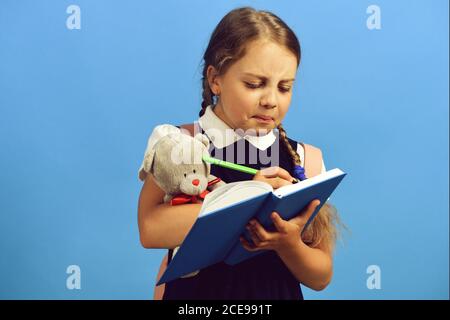 Schüler in Schuluniform mit Zöpfen und Rucksack. Mädchen hält Teddybär und schreibt in blauem Notizbuch mit Marker. Zurück zur Schule Konzept. Schulmädchen mit unzufriedenen Gesicht isoliert auf blauem Hintergrund Stockfoto