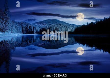 Bergsee unter dem Wald in der Nacht. Bäume in bunten Laub. Schöne Landschaft im Herbst Vollmondlicht. Wolken und Himmel spiegeln sich in der wa Stockfoto