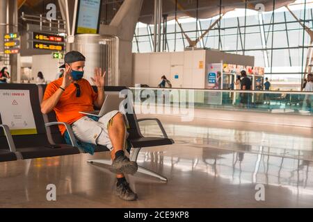 Junger Mann trägt eine Gesichtsmaske im Flughafen und Teilnahme an einem Online-Meeting auf seinem Laptop Stockfoto