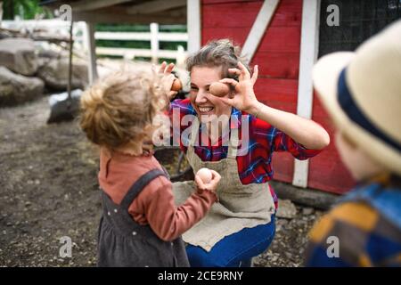 Fröhliche Mutter mit kleinen Kindern, die auf dem Bauernhof stehen, Eier sammeln. Stockfoto