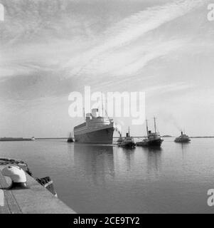 Historische Schlepper der 1950er Jahre ziehen den RMS Queen Elizabeth Ocean Liner in die ruhigen Gewässer des französischen Hafens von Cherbourg. Benannt nach der britischen Queen Mother, war es das größte Passagierschiff, das zu dieser Zeit (1938) gebaut wurde und blieb es 56 Jahre später. Sie wurde während des 2. Weltkriegs als Kriegsschiff eingesetzt und wurde erst 1946 wie beabsichtigt zum Ozeandampfer. Von da an bot dieser Luxusliner eine wöchentliche Verbindung zwischen Southampton, Großbritannien und New York City in den USA, über Cherbourg, Frankreich an. Im Jahr 1972 in Victoria Harbour, Hong Kong, während einer Umrüstung, fing sie Feuer und sank. Stockfoto