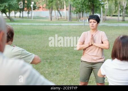 Fokussierter Yogalehrer, der Namaste Geste macht, während er mit Schülern meditiert Im Sommer Park Stockfoto