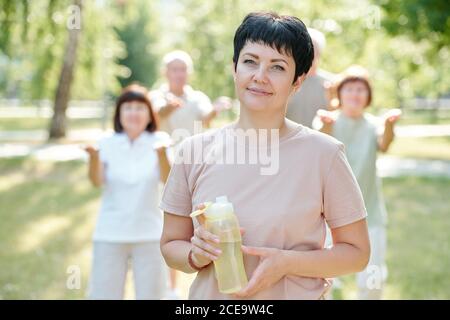 Portrait von lächelnden Brünette Fitness-Trainer mit kurzen Haaren halten Wasserflasche gegen ältere Menschen, die Sport treiben Stockfoto