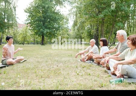 Kurzhaarige Yogalehrerin, die in Lotusposition sitzt und die Hände streicht Während des Gesprächs mit Yoga-Studenten vor dem Unterricht Stockfoto