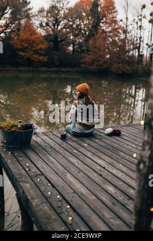 Rückansicht einer hübschen Frau, die auf einer hölzernen Veranda am Teich sitzt und im Herbst strickt. Stockfoto