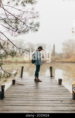 Seitenansicht des Fotografen, der mit der Kamera auf dem hölzernen Pier am Teich steht. Stockfoto