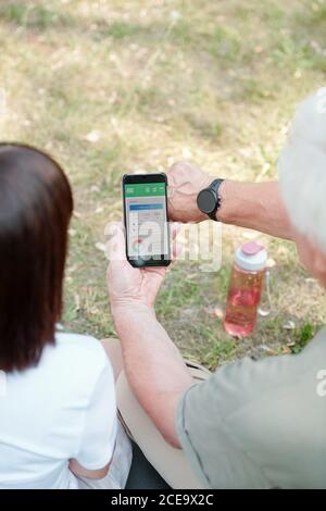 Über der Schulter Ansicht von Senior Mann synchronisiert Fitness-Tracker mit Smartphone beim Ausruhen mit Freundin nach dem Training Stockfoto