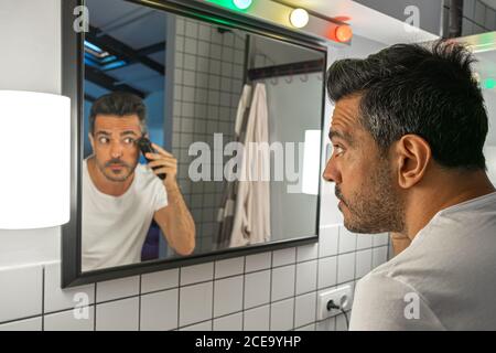 Der gutaussehende bärtige Mann rasiert sich mit der Trimmermaschine vor dem Badezimmerspiegel Stockfoto