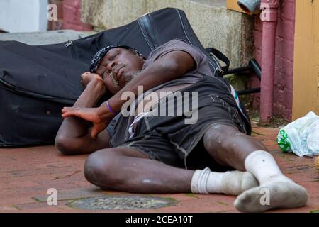 Annapolis, MD 08/21/2020: Ein obdachloser afroamerikanischer Mann schläft auf dem gepflasterten Bürgersteig auf seinem Gepäck. Seine Kleider sind Dorn aus und schmutzig. Stockfoto