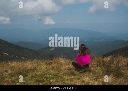 Rückansicht einer jungen Frau in zwanglosem Outfit, die auf einem grasbewachsenen Hügel sitzt und am bewölkten Tag in Bulgarien, Balkan, einen atemberaubenden Blick auf die wunderschöne Natur genießt Stockfoto