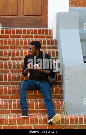 Annapolis, MD 08/21/2020: Ein junger afroamerikanischer Mann, der ein schwarzes T-Shirt, Jeans, Turnschuhe und ein Rucksack trägt, sitzt auf einer gemauerten Treppe draußen Stockfoto