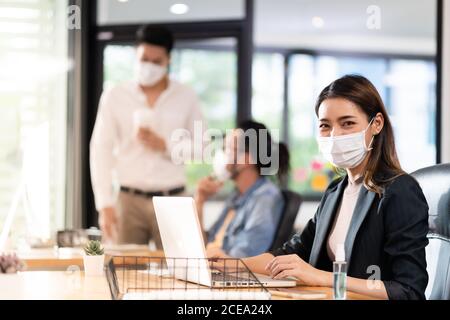 Portrait der asiatischen Büroangestellte Geschäftsfrau tragen Schutzmaske Arbeiten Sie in einem neuen normalen Büro mit interracial Kollegen im Hintergrund Als sozial Stockfoto