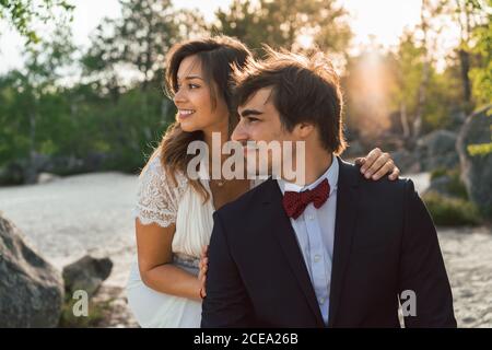 Fröhlicher, eleganter Mann und Frau in Hochzeitsoutfits umarmen sich am Strand und schauen lächelnd im Sonnenlicht weg Stockfoto