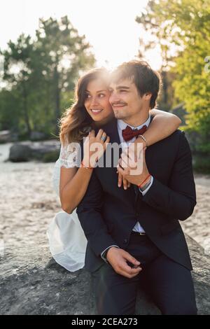 Fröhlicher, eleganter Mann und Frau in Hochzeitsoutfits umarmen sich am Strand und schauen lächelnd im Sonnenlicht weg Stockfoto