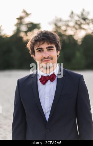 Erwachsene Mann trägt eleganten schwarzen Anzug mit Fliege und Stehen am Strand mit den Händen in den Taschen Blick auf die Kamera Stockfoto