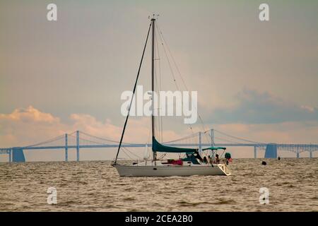 Bild eines Segelbootes, das über die Chesapeake Bay fährt, mit der Silhouette der berühmten Bay Bridge im Hintergrund. Es gibt Leute auf dem Boot, die sind Stockfoto