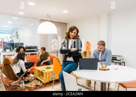Attraktive junge Frau, die auf dem Tisch sitzt und mit Kollegen im Co-Working-Büro zusammenarbeitet und die Kamera anschaut Stockfoto