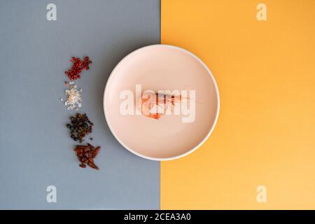 Haufen verschiedener Gewürze liegen neben der keramischen Platte mit klein Leckere Garnelen auf blauem und orangefarbenem Hintergrund Stockfoto