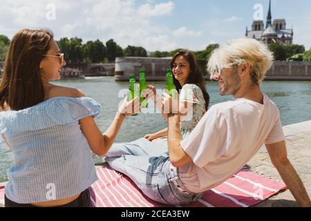 Seitenansicht von jungen Frauen in legerer Kleidung und stilvoll Mann, der sich am Wasser entspannt und zum Treffen trinkt Stockfoto