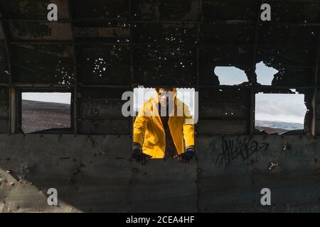 Hübscher junger Mann in gelber Jacke, der vor dem verlassenen Flugzeug steht Karkasse und Blick auf die Kamera während der Reise durch Island Stockfoto