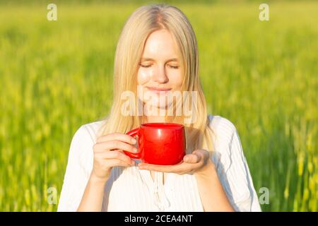 Schöne junge Frau, die an einem sonnigen Sommertag in der Natur ein Getränk trinkt. Grüner Feldhintergrund. Tee/Kaffee. Stockfoto