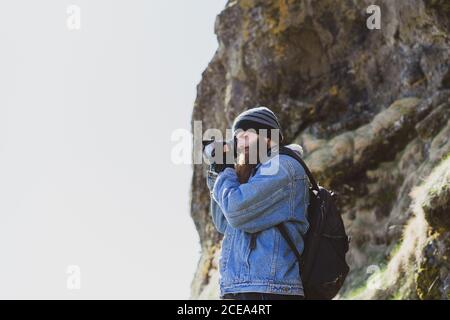 Seitenansicht des bärtigen Mannes im Hut mit Rucksack nehmen Bild mit Kamera von der isländischen Landschaft Stockfoto