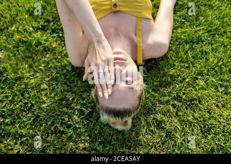 Crop-Ansicht der schönen Frau in gelben Sommerkleidung liegen Auf sonnenbeschienenen Rasen und die Hälfte des Gesichts mit der Hand bedeckend Blick auf die Kamera von oben Stockfoto