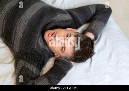 Von oben Schuss des schönen jungen Mannes im Pullover liegend Auf dem Bett mit Händen hinter dem Kopf und Blick auf die Kamera Stockfoto