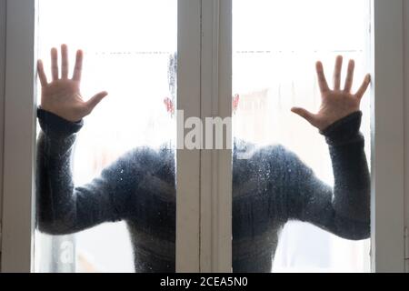 Aufnahme eines Mannes im Pullover, der hinter nassem, dampfendem Glas steht Die Tür hält die Hände darauf Stockfoto