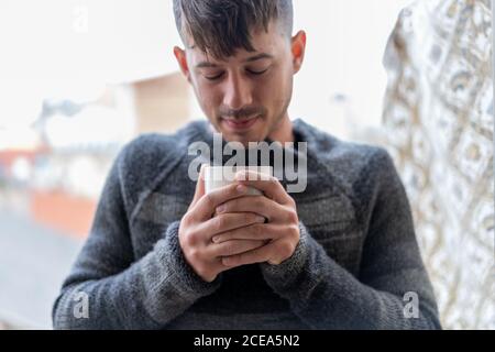 Junger Mann in grau kuschelig Pullover stehend mit Tasse Kaffee genießen Geruch und Wärme Stockfoto