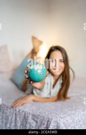 Verschwommene junge Frau, die zu Hause auf einem bequemen Bett liegt, einen kleinen Globus vor die Kamera zeigt Stockfoto