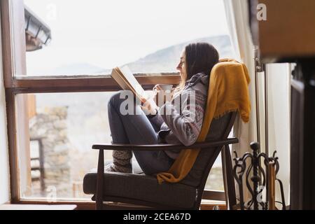 Seitenansicht der jungen Dame mit Tasse frisch heiß Trinken Sie beim Lesen des interessanten Buches, während Sie neben dem riesigen Fenster sitzen Gemütliches Zimmer Stockfoto