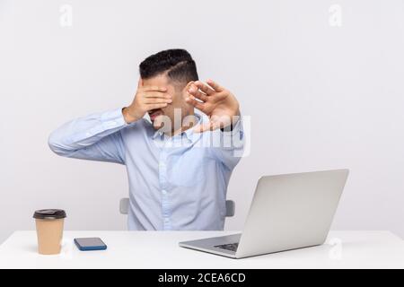 Nein, ich will nicht hinsehen! Geschäftsmann sitzt Büroarbeitsplatz mit Laptop auf dem Schreibtisch, die Augen zeigen Stop-Geste, vermeiden sehen etwas unpleas Stockfoto