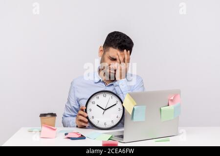 Überstunden. Unglücklicher, trauriger Mann, der am Büroarbeitsplatz sitzt, die Uhr hält und die Facepalm-Geste hält und Bedauern über die verpasste dea zum Ausdruck bringt Stockfoto